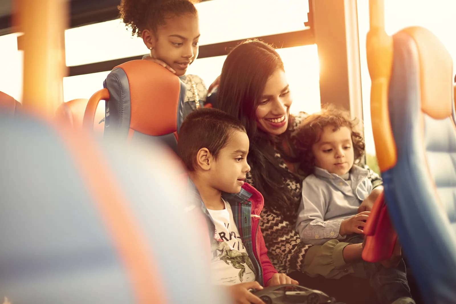 A smiling mother rides a bus with her three young children, one girl leaning on a seat in front and two boys sitting next to their mother.