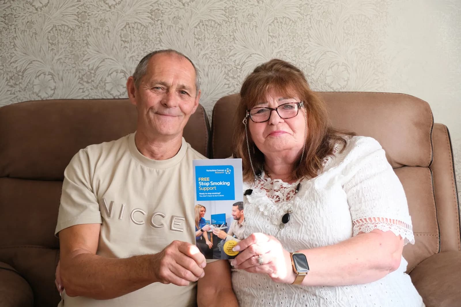 A man and a woman sitting on brown armchairs hold up a leaflet that says 'Free Stop Smoking Support'.