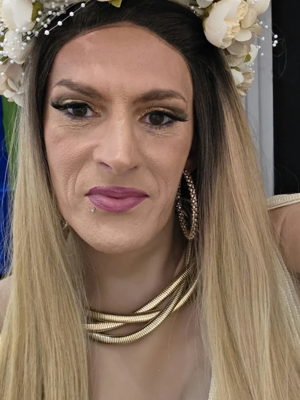 A person with long blonde hair, a floral headpiece, gold hoop earrings, a gold coiled necklace, and bright makeup including pink lipstick and golden eyeshadow gazes directly at the camera.