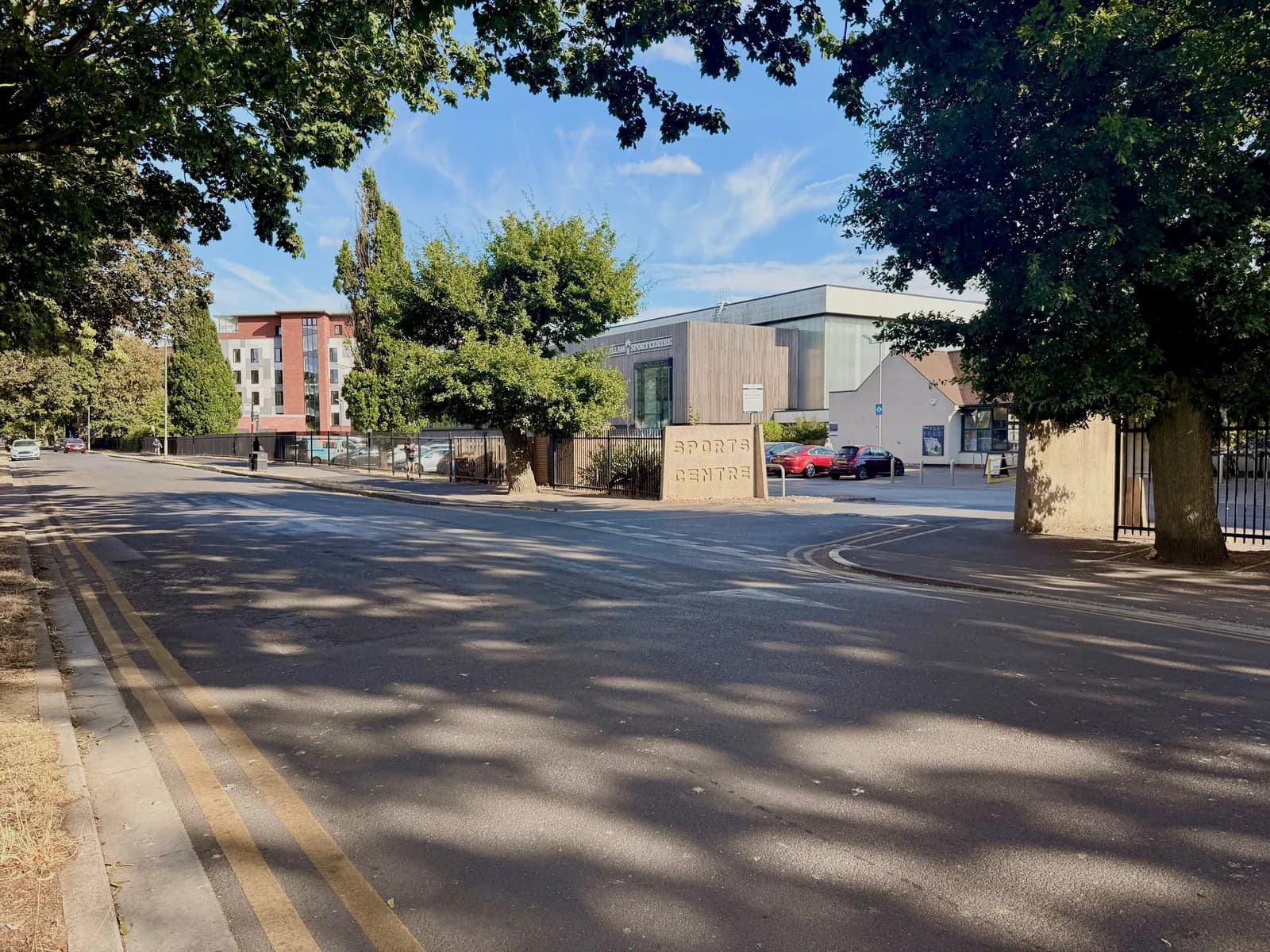 A sports center sign stands at the entrance of a modern building complex with cars parked outside, framed by an empty road and green trees under a bright sky.