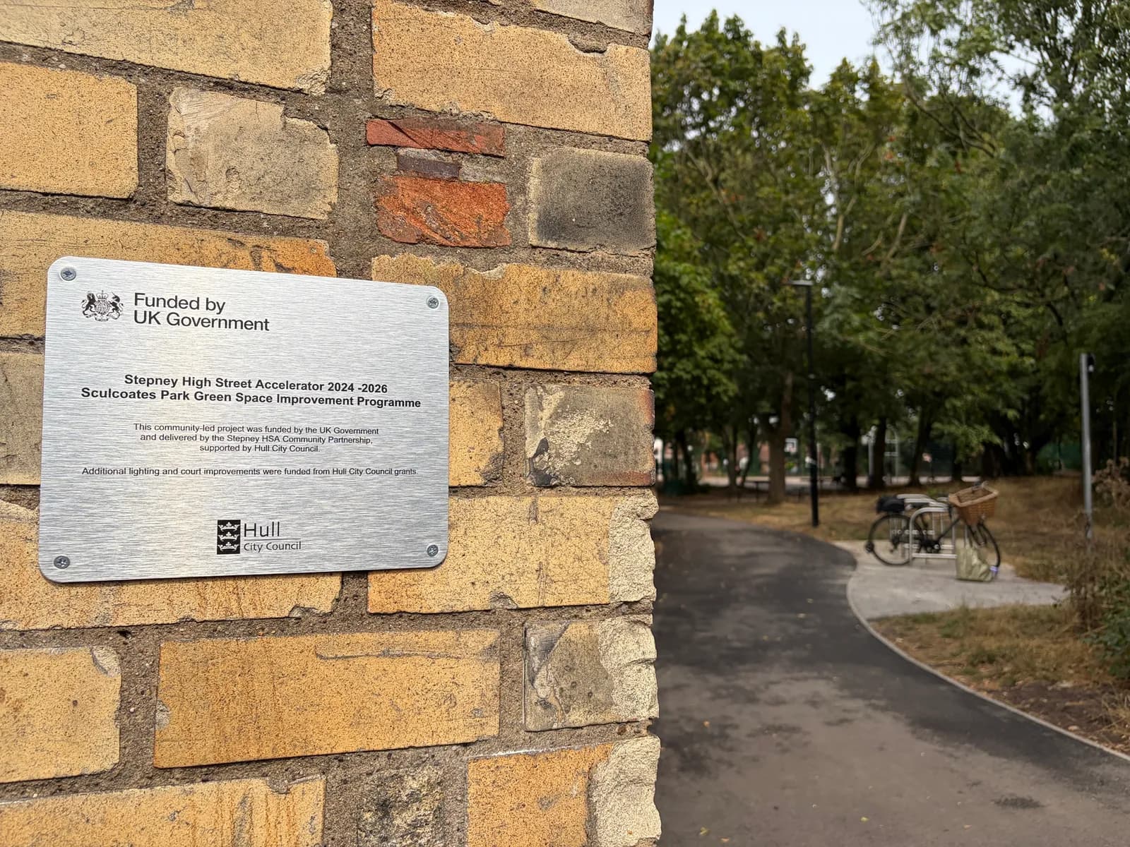 A plaque on a brick wall explains that the Stepney High Street Accelerator 2024-2026 Sculcoates Park Green Space Improvement Programme is funded by the UK Government and Hull City Council, with a park path and bicycle visible in the background.