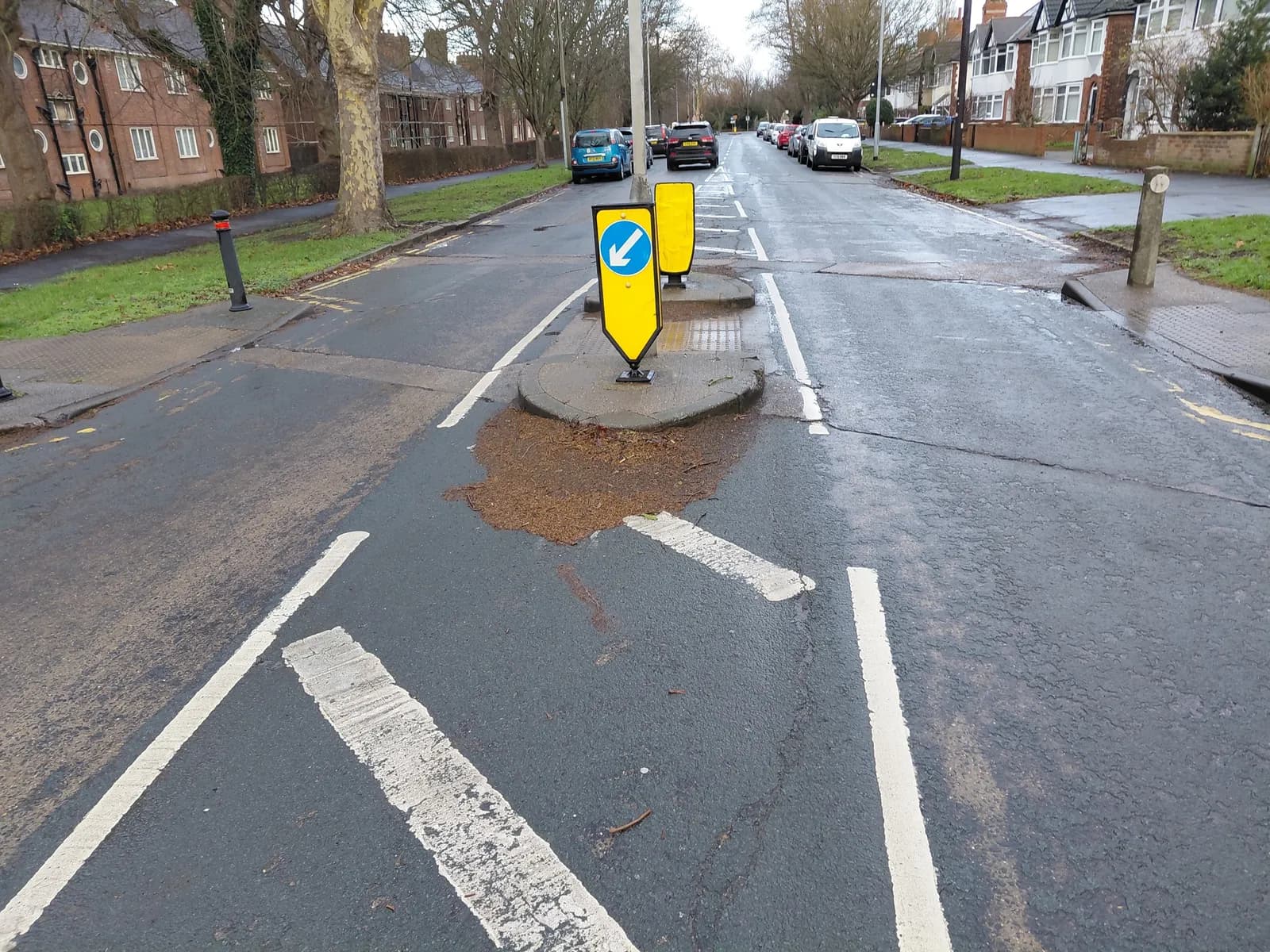 A yellow and blue arrow sign on a traffic island in the middle of a wet asphalt road, with parked cars and residential buildings in the background.