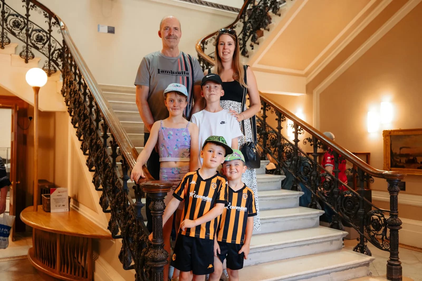A family of six, including two adults and four children, stands smiling on a grand marble staircase with ornate railings.