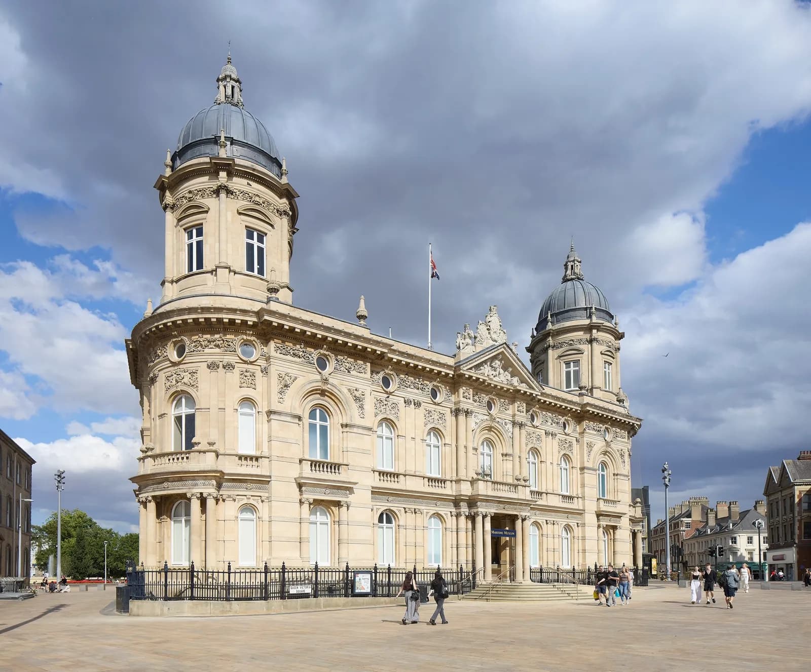 A grand, ornate beige stone building with two large domed towers stands under a cloudy sky, with people walking on the paved square in front of it.